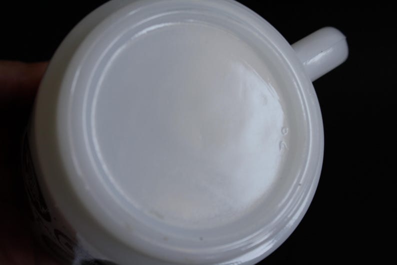 May include: A white ceramic mug with a rounded base and a small handle. The bottom of the mug has a slightly recessed center and a raised rim. The number "26" is imprinted on the bottom.