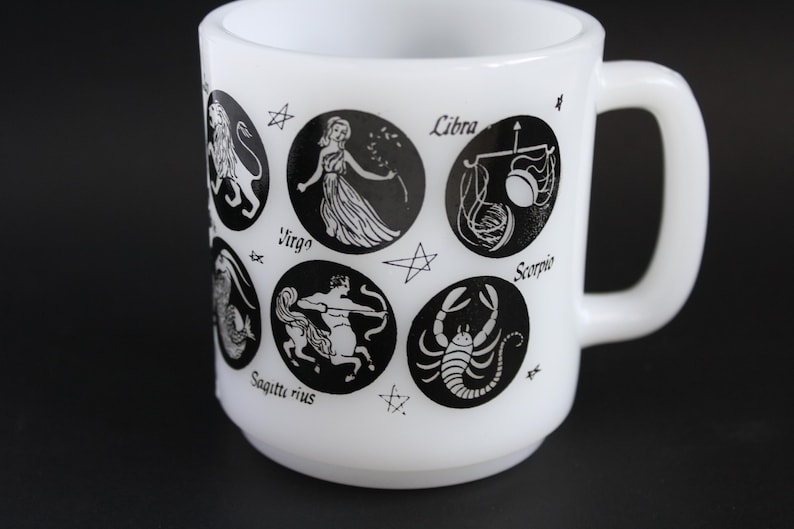 May include: White ceramic mug featuring black zodiac symbols and names. The mug has a rounded body and a large handle. Zodiac signs include Virgo, Libra, and Scorpio. A unique gift for astrology enthusiasts.