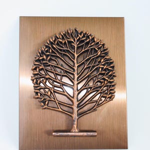 May include: A copper-coloured metal wall art piece featuring a stylised tree with intricate branches and leaves. The tree is mounted on a square copper-coloured metal background.