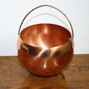 May include: A copper bowl with a brass handle and three legs. The bowl is round and has a shiny finish.