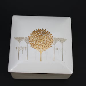 May include: White square ceramic box with a gold and brown tree design. The tree is in the centre of the box and has leaves that are a light brown colour. The background of the box is white.