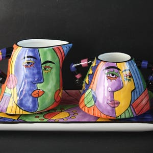 May include: A colorful ceramic creamer and sugar bowl set with a matching tray. The set features abstract face designs in blue, green, purple, and yellow. The handles are striped. The tray is rectangular with a white border.