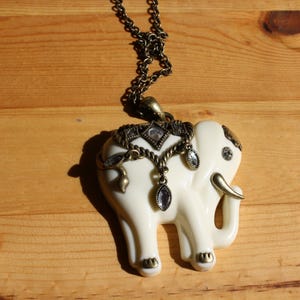 May include: Ivory-colored elephant pendant necklace with antique gold-tone accents. The elephant pendant features ornate detailing and dangling teardrop-shaped gems. The necklace chain is a dark, antique gold color.