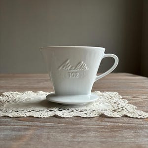 Puede incluir: Filtro de café de cerámica blanca con el texto "Melitta 102" en relieve en el lateral. El filtro está sobre un encaje blanco.