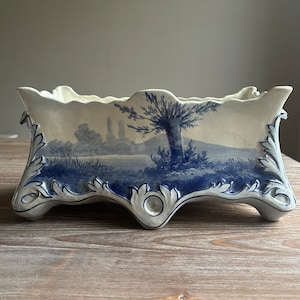 Peut inclure: Un pot en céramique blanche avec un motif de paysage bleu et blanc. Le pot a un bord festonné et quatre pieds.
