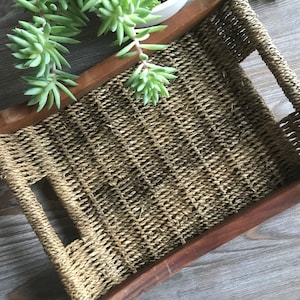 May include: A woven tray with wooden sides and two cut-out handles. The tray is made of natural brown rope and has a rustic look.