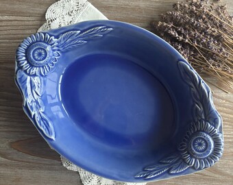 May include: A blue ceramic baking dish with a white sunflower design. The dish is oval shaped and has a raised rim.