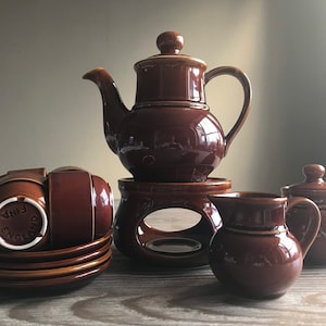 May include: A brown ceramic tea set with a teapot, creamer, sugar bowl, four cups, and four saucers. The set is on a wooden table and includes a stand for the teapot.