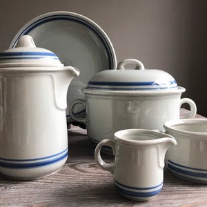 Può includere: Un set di stoviglie da cucina in ceramica bianca con strisce blu. Il set comprende una brocca per la panna, una zuccheriera, una casseruola con coperchio e un piatto da portata.