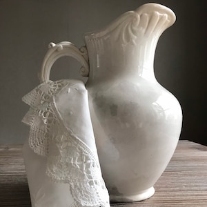 May include: A white ceramic jug with an ornate handle and a delicate lace doily draped over the spout. The jug is sitting on a wooden surface.