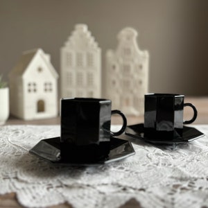Peut inclure: Deux tasses à café en céramique noire avec des soucoupes assorties sur une nappe en dentelle blanche. Les tasses ont une forme géométrique et sont nettes. L'arrière-plan présente des maisons en céramique blanche.