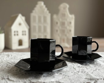 May include: Two black ceramic coffee cups with matching saucers on a white lace tablecloth. The cups have a geometric shape and are in focus. The background features white ceramic houses.