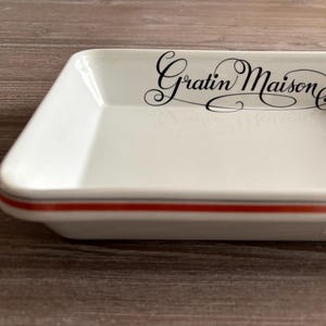 Può includere: Piatto da forno rettangolare bianco con un bordo rosso e grigio. Il piatto ha le parole "Gratin Maison" stampate in corsivo nero sul davanti.
