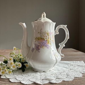 Op de afbeelding: Witte porseleinen theepot met een bloemenmotief met paarse en roze bloemen. De theepot heeft een gouden rand rond de handgreep en de tuit.
