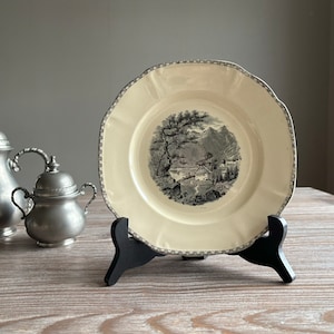 May include: A black and white ceramic plate with a decorative border and a scene of a mountain landscape with a small house. The plate is on a black stand.