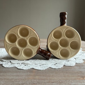 Puede incluir: Dos ramequines de cerámica marrón y beige con asas. Cada ramequín tiene siete hendiduras individuales para cocinar pequeñas porciones de comida.