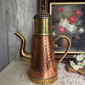 Puede incluir: Una cafetera de cobre y latón con textura martillada. La cafetera tiene un pico curvo y un asa. La tapa y el asa son de latón. La cafetera está sobre una superficie blanca.
