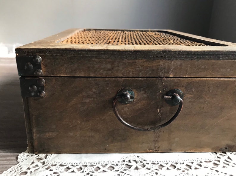 Puede incluir: Una caja de madera con una tapa tejida y una manija de metal. La caja es marr&oacute;n y tiene detalles met&aacute;licos.