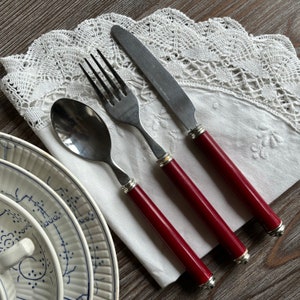 Puede incluir: Un juego de tres cubiertos plateados con mangos rojos. El juego incluye un tenedor, un cuchillo y una cuchara. Los cubiertos están dispuestos sobre un paño blanco con un borde de encaje.