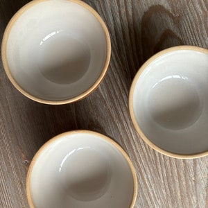 May include: Three white ceramic bowls with a light brown rim. The bowls are sitting on a light brown wooden surface.