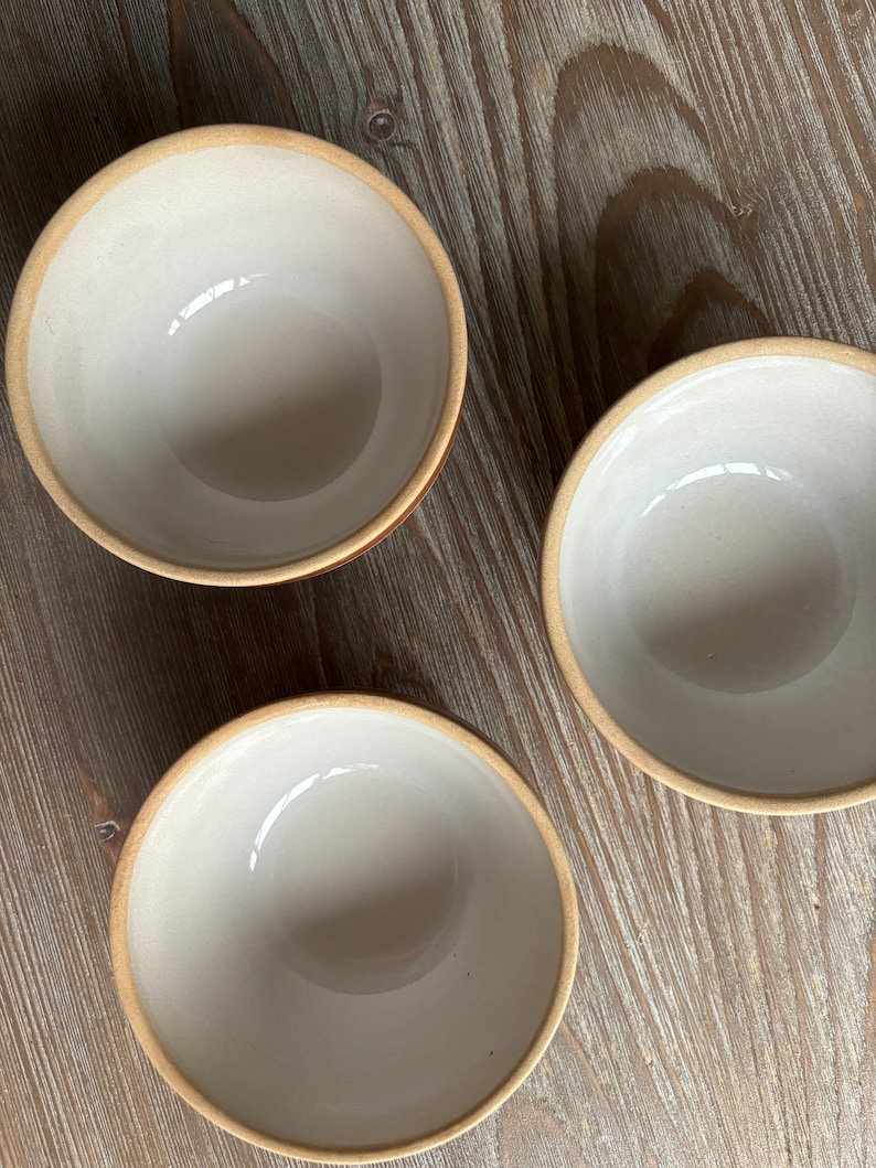 May include: Three white ceramic bowls with a light brown rim. The bowls are sitting on a light brown wooden surface.