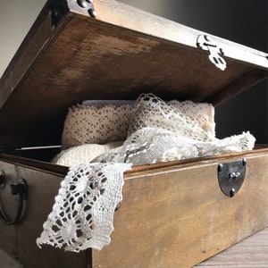Puede incluir: Una caja de madera con tapa abatible y detalles met&aacute;licos. La caja est&aacute; abierta y contiene encaje blanco.
