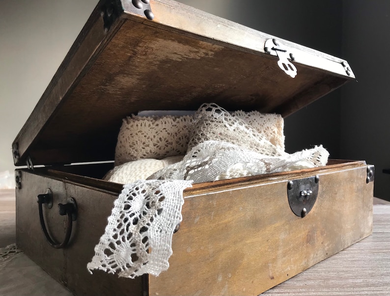 Puede incluir: Una caja de madera con tapa abatible y detalles met&aacute;licos. La caja est&aacute; abierta y contiene encaje blanco.