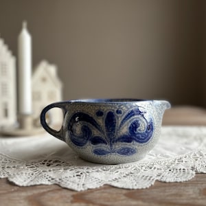 Könnte beinhalten: Eine blaue und weiße Keramik-Sahnegießer mit einem floralen Design. Der Sahnegießer hat einen Griff und einen Ausguss. Er steht auf einer weißen Spitzenunterlage.