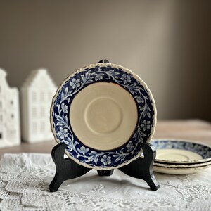 May include: A blue and white porcelain saucer with a gold rim and floral pattern. The saucer is on a black stand and a white lace tablecloth.