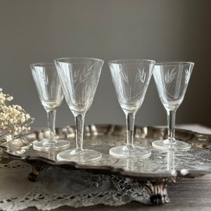 May include: Four clear, stemmed glasses with etched wheat stalk designs are arranged on a silver serving tray. The glasses have a delicate, vintage aesthetic. The tray has ornate edges and sits on decorative feet. A sprig of white flowers is in the foreground.