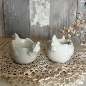 Op de afbeelding: Twee witte keramische eierrekjes in de vorm van kippen op een gehaakt onderzetter. De rekjes zijn gevormd als kippen met hun kop naar beneden gekanteld.