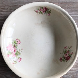 Könnte beinhalten: Eine weiße Keramikschüssel mit einem Blumendesign mit rosa Rosen und grünen Blättern. Die Schüssel hat eine leicht abgenutzte Optik.