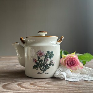 Op de afbeelding: Witte theepot van keramiek met een bruin deksel en handvat. De theepot heeft een bloemenmotief met roze en groene bloemen en de tekst "Alcea rosea".