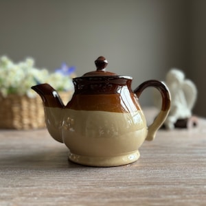 Puede incluir: Una tetera de cerámica marrón y beige con cuerpo redondeado y un pico. La tetera tiene una tapa marrón con un pomo en la parte superior.