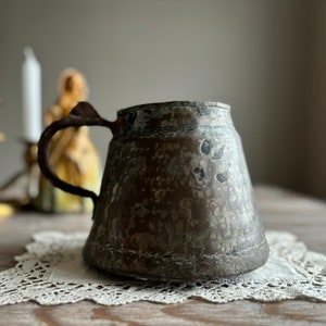 Könnte beinhalten: Ein antiker Kupferkrug mit einer abgenutzten, rustikalen Oberfläche. Der Krug hat einen Henkel und einen breiten, abgerundeten Boden. Die Oberfläche des Kruges ist mit einer Patina des Alters bedeckt.