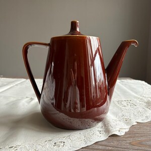 Peut inclure: Une théière en céramique brune avec un bec large et une poignée. La théière a une finition brillante et une forme arrondie.