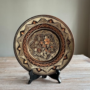Può includere: Un piatto decorativo in rame con intricati disegni floreali in oro e rame. Il piatto è su un supporto nero.