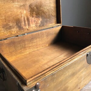 Puede incluir: Una caja de madera vac&iacute;a con una tapa con bisagras y detalles met&aacute;licos. La caja est&aacute; hecha de madera marr&oacute;n claro y tiene un acabado natural. La caja est&aacute; abierta y el interior es visible.