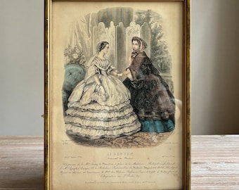 May include: Framed vintage print featuring two women in elaborate dresses, holding hands. The print, titled "LE BON TON Journal de Modes," is in a gold-colored frame. The women are set against a backdrop of a decorative screen and a floral arrangement.