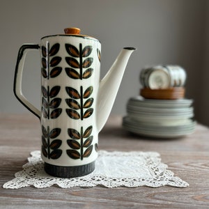 May include: A white ceramic teapot with a black and brown leaf pattern. The teapot has a gold lid and a spout. It is sitting on a white lace doily on a wooden table.