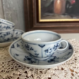 Könnte beinhalten: Ein weißes Tassen- und Untertassen-Set mit blauen Blumenmustern. Die Tasse hat einen kleinen Henkel und steht auf einer passenden Untertasse. Das Design zeigt zarte blaue Blumen und Ranken. Das Set steht auf einer cremefarbenen Spitzendecke.