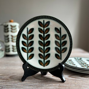 May include: A white ceramic plate with a green and brown leaf design. The plate is on a black stand.