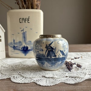 May include: A small blue and white ceramic jar with a silver lid. The jar features a Dutch windmill scene with a blue and white color scheme. The jar is sitting on a white lace doily.