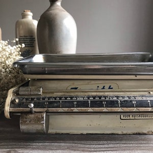 Op de afbeelding: Een vintage witte keukenweegschaal met een zilveren bovenkant en een zwarte schaal gemarkeerd in grammen. De weegschaal heeft een blauw label met de tekst "71:  VOOR HUISHOUDELIJK GEBRUIK".