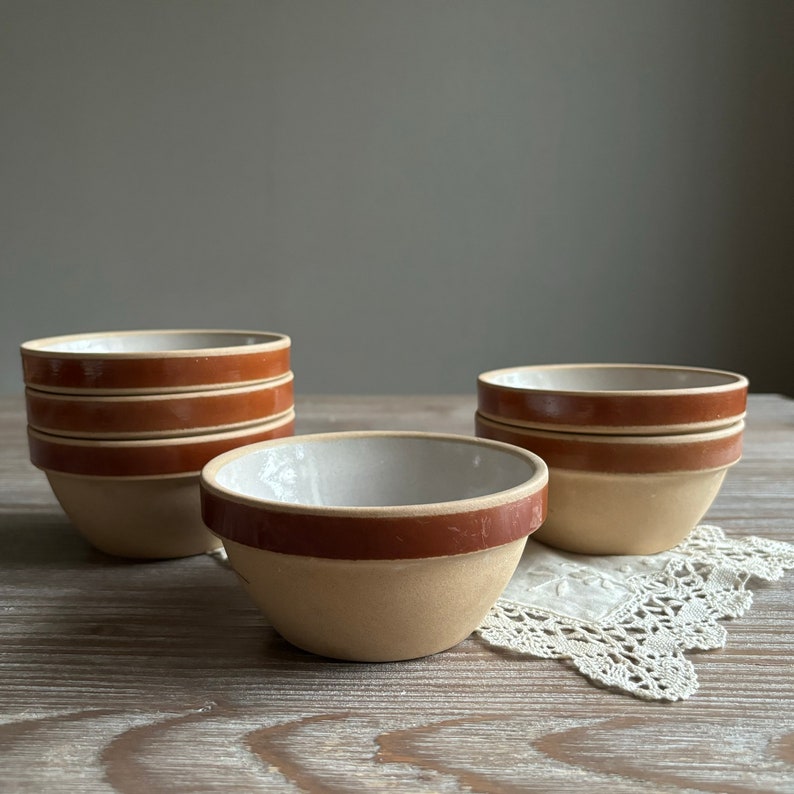 May include: A set of five small, beige ceramic bowls with a brown rim. The bowls are stacked on top of each other, with one bowl sitting on a white lace doily.