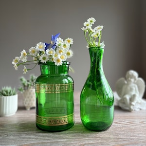 Può includere: Due vasi di vetro verde con finiture dorate, uno con un'apertura larga e l'altro con un collo stretto, entrambi pieni di fiori bianchi.