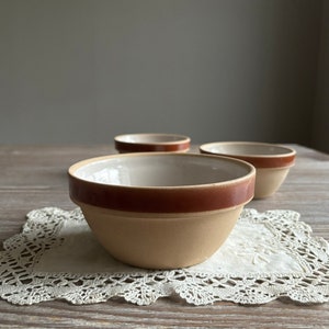 May include: Three small, beige ceramic bowls with brown rims. The bowls are sitting on a white crocheted doily on a wooden surface.