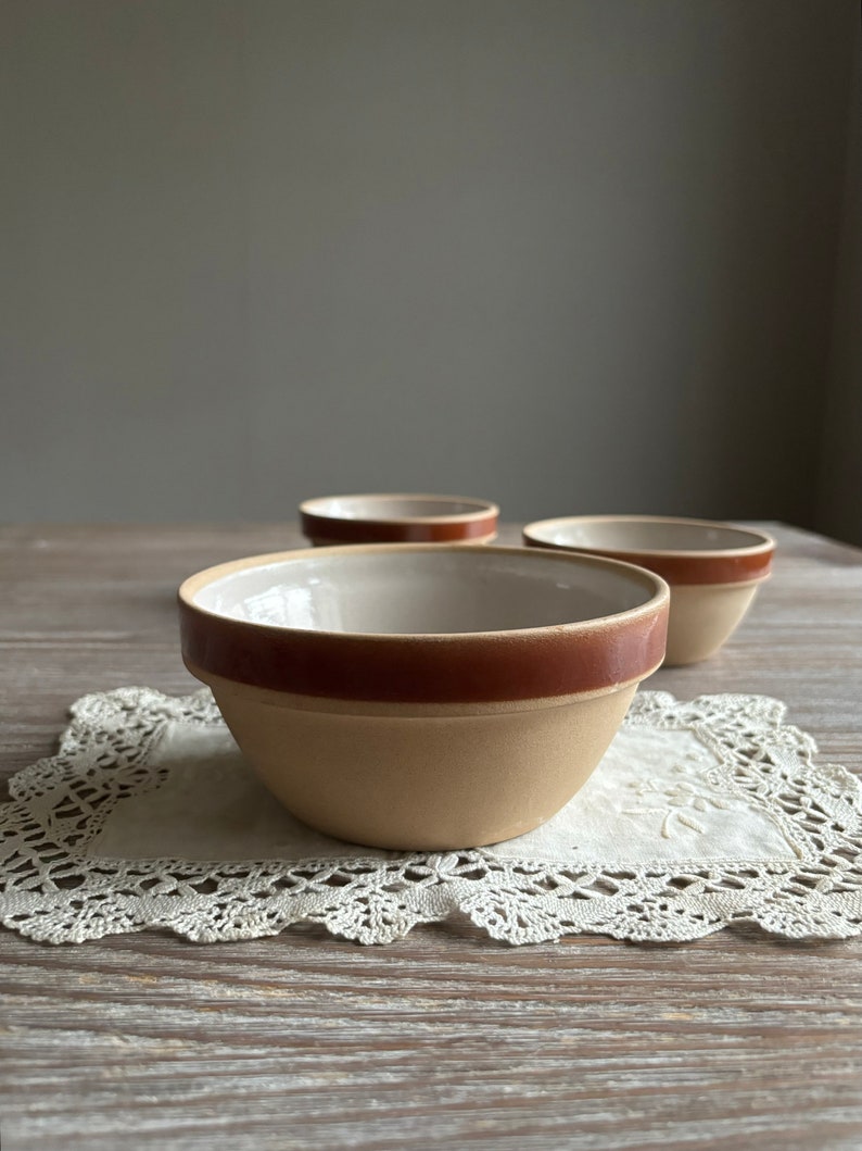 May include: Three small, beige ceramic bowls with brown rims. The bowls are sitting on a white crocheted doily on a wooden surface.