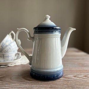 Op de afbeelding: Een witte theepot van keramiek met een blauw-wit ontwerp. De theepot heeft een rond lichaam en een tuit aan de zijkant. De deksel is ook blauw-wit.