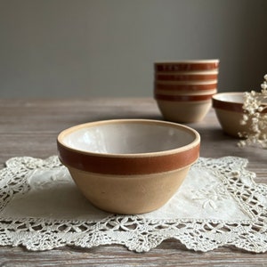 May include: A set of small, white ceramic bowls with a brown rim. The bowls are sitting on a white lace doily on a wooden table.
